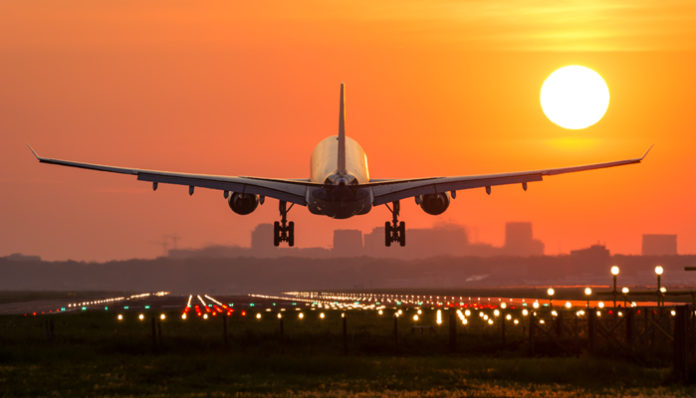 Image of a jet about to land on the runway. The sky is transitioning red, orange, yellow from left to right. A city is silhouetted in the distance.