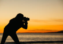 Silhouette of person taking photo near water, orange sunset background