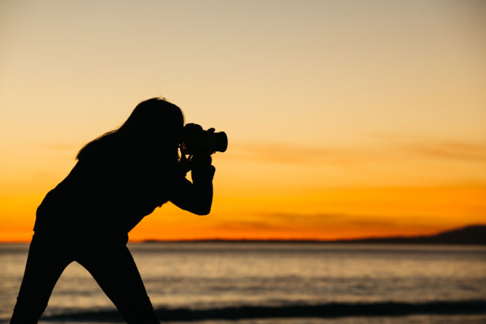 Silhouette of person taking photo near water, orange sunset background