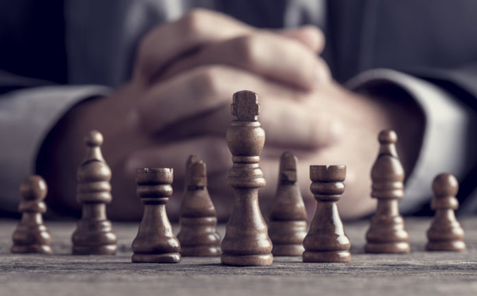 Image of a businessman's clasped hands and and chess pieces on an old wooden table. Photo by Gajus via Shutterstock license.